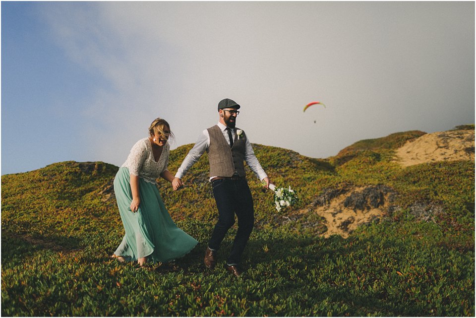 groom leads bride across a sand dune laughing with a hang glider in the background