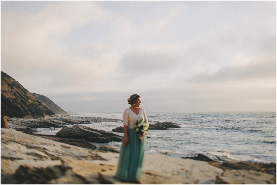 bride stands on rock next to the ocean and looks out to sea