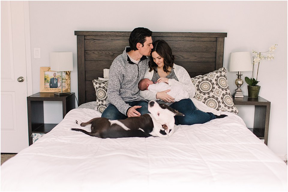 mother and father on bed holding newborn baby with the family dog lying by them