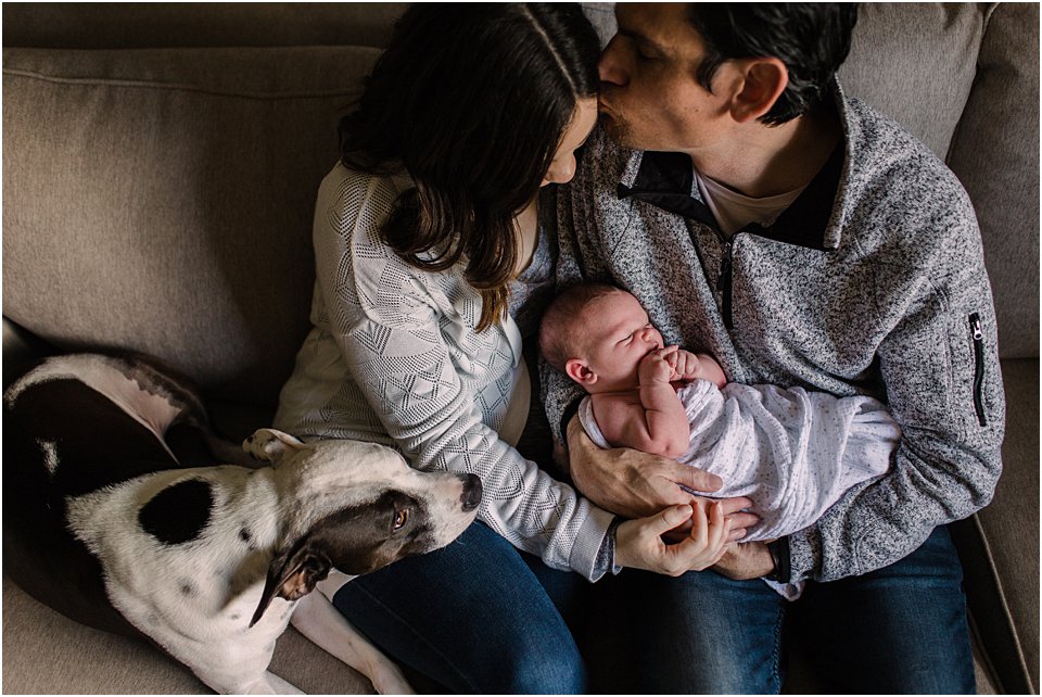 mother and father on the couch holding newborn baby with family dog