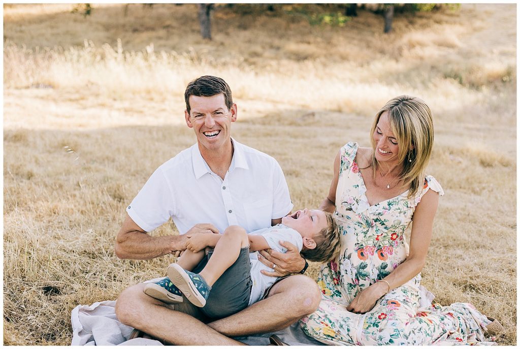family has tickle fight seated on blanket in golden field 